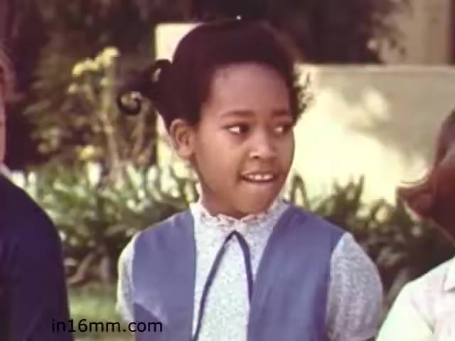 The still from 'Walk Safely,' an educational film from 1982 by Fiesta Films, features a young girl in a blue dress with a white collar, looking towards someone off-camera. Her expression suggests curiosity or intrigue. The background has greenery, typical of an outdoor setting, contributing to a light, engaging atmosphere suitable for educational content. The film likely addresses themes of safety and awareness in a child-friendly manner.