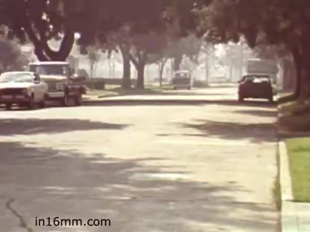The still from 'Walk Safely,' an educational film from 1982, depicts a quiet suburban street lined with trees. The scene is characterized by a hazy atmosphere, suggesting either early morning or late afternoon light. Vehicles, including a few cars and a truck, are parked along the side of the road. The setting feels calm and emphasizes a theme of safety, likely linked to pedestrian awareness in a residential area. The film likely aims to educate viewers about walking safely in such environments.