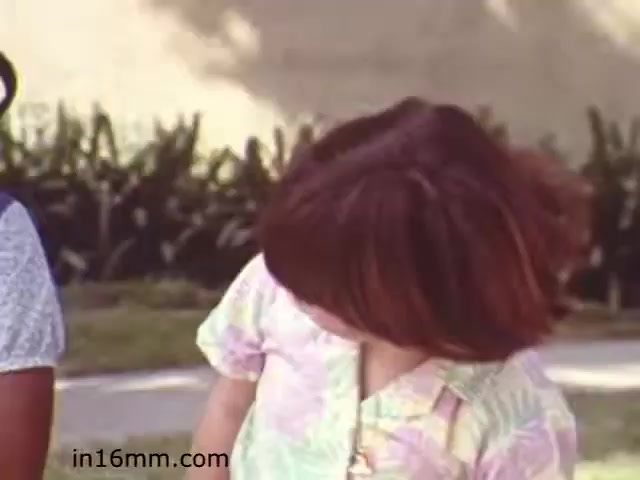 The still from 'Walk Safely,' an educational film produced by Fiesta Films in 1982, features two children in a casual outdoor setting. One child appears to be looking down, while the other is close by, possibly interacting with her. The clothing is colorful and patterned, reflective of the fashion trends of the early '80s. The background shows greenery, suggesting a suburban or park-like environment. This film likely aimed to educate children about safety while walking, using relatable scenarios to convey important messages.