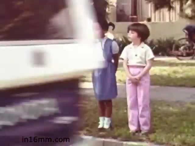 The still from 'Walk Safely,' an educational film from 1982 by Fiesta Films, depicts two children standing on a sidewalk, appearing to wait for a vehicle to pass. The background shows residential buildings and greenery. The scene emphasizes pedestrian safety, likely illustrating the importance of being cautious around traffic. The visual style is characteristic of 1980s educational films, featuring bright colors and a simple composition.