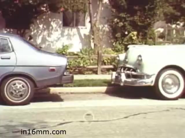 The still from 'Walk Safely,' an educational film from 1982 by Fiesta Films, features two parked cars on a suburban street. On the left is a blue vehicle, and on the right is a white car with a distinctive front grill. The setting appears to be in a residential area, with green foliage and a home visible in the background. This type of scene likely aims to highlight street safety and the importance of being aware of one's surroundings when walking near vehicles.