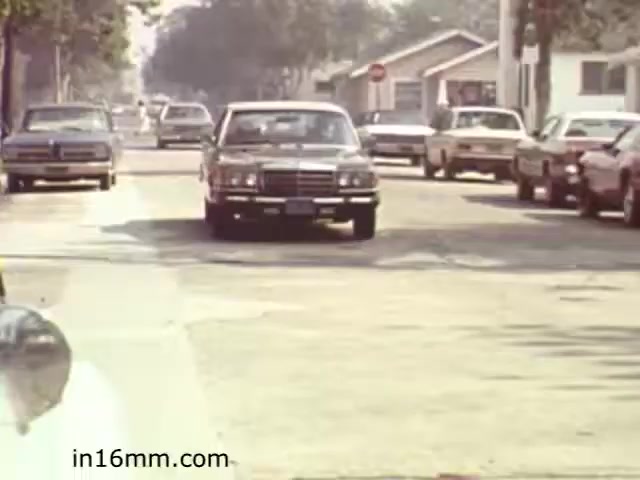 The still from 'Walk Safely,' an educational film from 1982 by Fiesta Films, likely captures a typical street scene from that era. In the image, several cars are parked along a residential street, while a vehicle drives down the road. The overall atmosphere suggests a focus on urban or suburban safety, possibly emphasizing pedestrian awareness and traffic rules. The aesthetic reflects the 1980s style, characterized by the design of the vehicles and the casual look of the surroundings.