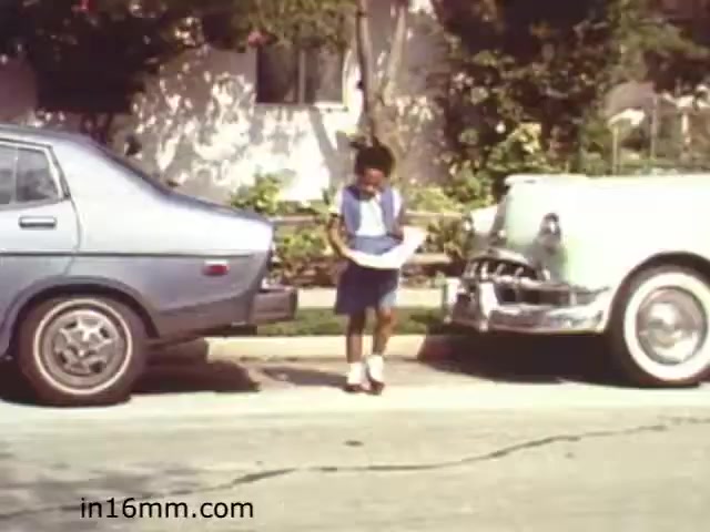 The still from the 1982 educational film 'Walk Safely' shows a young child standing between two parked cars on a residential street. The child appears to be holding a piece of paper, possibly a map or instructions. The scene conveys a focus on pedestrian safety, highlighting the importance of awareness in urban environments. The backdrop features a suburban setting with greenery and a house, characteristic of the time period.