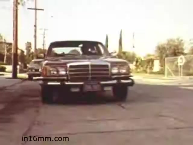The still from the 1982 educational film 'Walk Safely' by Fiesta Films depicts a vintage car approaching down a suburban street. The scene suggests a focus on pedestrian safety and road awareness, likely illustrating the relationship between vehicles and pedestrians, a common theme in educational films. The surroundings feature utility poles and a few trees, typical of a residential area. The image captures a moment that likely serves as a visual aid for teaching safe walking practices.