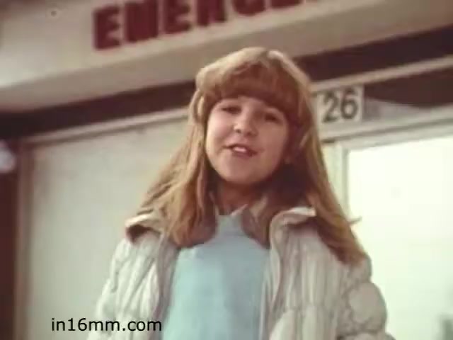 The still from 'Walk Safely,' an educational film from 1982 produced by Fiesta Films, features a young girl with blonde hair, wearing a light colored sweater and a white puffy jacket. She appears to be speaking or presenting something, possibly conveying a safety message related to walking or pedestrian awareness. The background shows a building with the word 'EMERGENCY' visible, suggesting a location relevant to health or safety. The overall tone seems educational and engaging, primarily aimed at children.