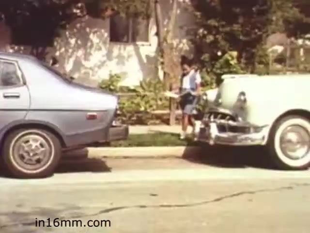 The still from 'Walk Safely' depicts a child standing on a sidewalk between two parked cars, a blue sedan and a vintage white vehicle. The setting appears suburban, with greenery and houses in the background. The child is holding something in their hands, likely related to pedestrian safety, which aligns with the educational theme of the film. The scene captures a moment emphasizing safety awareness, particularly for children in urban environments. The image reflects the style and tone typical of educational films from the early 1980s.