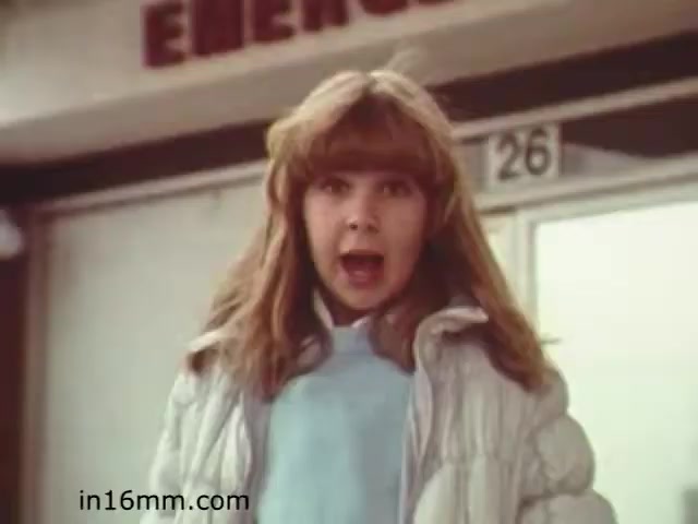 The still from 'Walk Safely' features a young girl with long, blonde hair, wearing a white puffy jacket and a light blue top. She appears to be expressing surprise or concern, possibly emphasizing a safety message. The background includes a sign that reads 'EMERGENCY.' The film, produced in 1982 by Fiesta Films, likely aims to educate about safety, particularly for children. The overall tone seems urgent, highlighting the importance of being aware of one's surroundings.