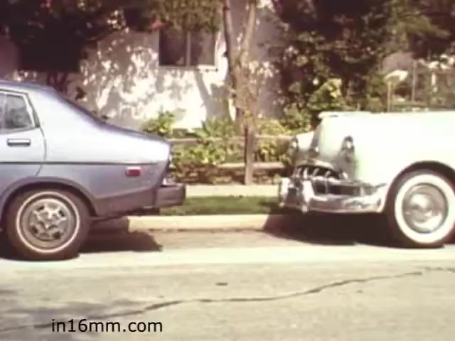The still from 'Walk Safely - Educational Film' shows two parked cars on a street: a blue sedan on the left and a white vintage car on the right. The scene appears to be set in a suburban area with greenery in the background, suggesting a focus on pedestrian safety and traffic awareness. The film likely aims to educate viewers about road safety or safe walking practices in urban environments.