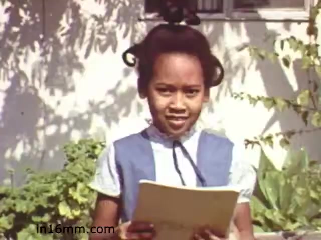 The 16mm still from 'Walk Safely,' an educational film from 1982 produced by Fiesta Films, features a young girl standing outdoors. She has curly hair styled in a bun and is wearing a blue dress with a white collar. The background includes green foliage, suggesting a sunny, inviting setting. The girl appears to be reading from a booklet or script, likely delivering a message related to pedestrian safety, which is the theme of the film.