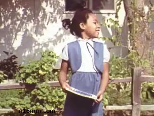 The still from 'Walk Safely,' an educational film from 1982, features a young girl standing outdoors. She is dressed in a blue outfit with a white top and holds a book or folder in her hands. The background includes a wooden fence and greenery, suggesting a residential or community setting. The girl appears to be smiling or looking thoughtfully, capturing a moment that likely emphasizes themes of safety and awareness in the context of childhood.