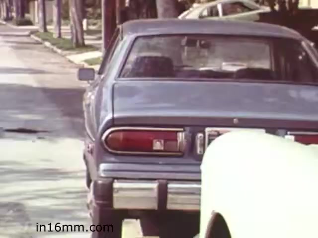 The still from 'Walk Safely,' an educational film from 1982 produced by Fiesta Films, depicts a suburban street scene featuring two parked cars. The focus is on a gray vehicle with a prominent rear view, displaying its taillights and bumper. The street appears lined with trees and other parked cars, creating a typical residential atmosphere. This film likely aimed to convey messages about pedestrian safety, and this scene may set the context for discussing safe walking practices in urban environments.