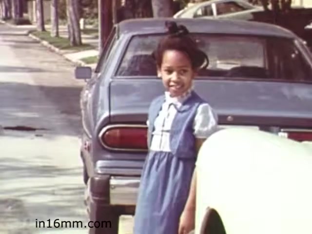 The still from the 1982 educational film 'Walk Safely' by Fiesta Films features a young girl wearing a blue dress with a white collar, standing near the back of a parked car on a suburban street. She has a playful expression and appears to be engaged in the moment. The street is lined with trees and other parked cars, evoking a safe, residential environment. The film likely aimed to teach children about pedestrian safety in a relatable context.