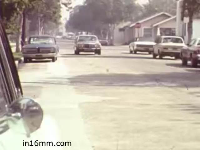 The still from 'Walk Safely,' an educational film from 1982 by Fiesta Films, depicts a street scene typical of a suburban area during that time. The focus appears to be on the traffic and road conditions, highlighting vehicles parked along the curb and one car driving down the street. The environment is well-lit and features trees lining the street, suggesting a safe and inviting neighborhood setting. This frame likely serves to illustrate pedestrian safety or traffic awareness.