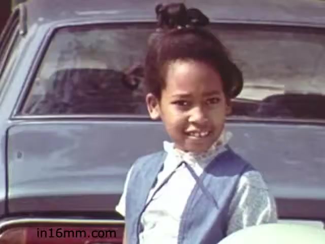 The still from 'Walk Safely,' an educational film from 1982 by Fiesta Films, features a young girl with a confident expression, dressed in a blue vest over a white collared shirt. She stands in front of a car, suggesting a casual, outdoor setting. The image aims to convey themes of safety and awareness, typical of educational films from that era, which often targeted children to encourage safe practices in public spaces.