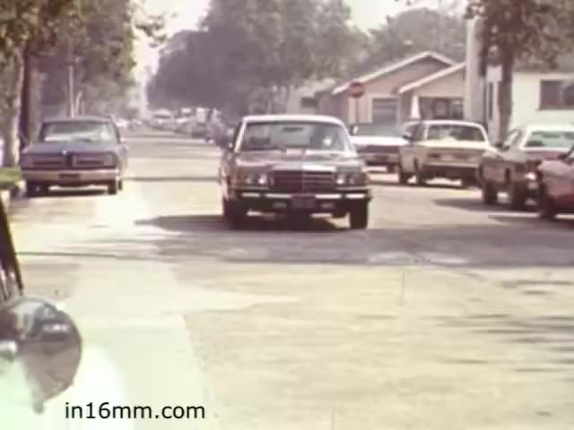 The still from 'Walk Safely' features a quiet street scene typical of 1982, showcasing several parked cars and a couple of moving vehicles. The focus is on promoting pedestrian safety, likely highlighting the importance of being aware of traffic while crossing the street. The suburban setting includes houses on either side, with trees lining the street, reflecting the era's aesthetic. Overall, it serves as a reminder for viewers to be cautious in their surroundings.