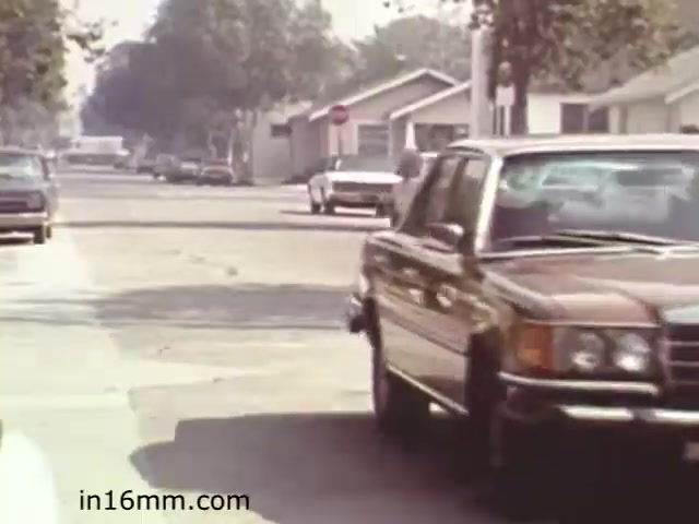 The still from 'Walk Safely,' an educational film from 1982 by Fiesta Films, depicts a suburban street scene. It features a brown car driving past on a residential road lined with trees and houses. In the background, additional vehicles are parked along the street, and a stop sign can be seen. The overall atmosphere suggests a focus on safety, possibly addressing pedestrian awareness or traffic rules in suburban environments.