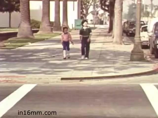 The still from 'Walk Safely,' an educational film from 1982 by Fiesta Films, features two children walking on a sidewalk, seemingly aware and cautious of their surroundings. The scene captures a suburban environment, with palm trees lining the street and parked cars visible. The children, dressed in casual attire, appear to be in a moment of learning about pedestrian safety. The setting emphasizes the importance of being aware while walking, fitting the film's educational theme.