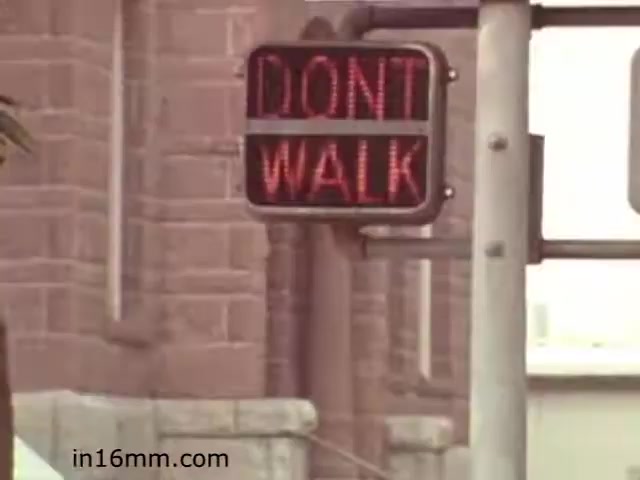 The still from the 1982 educational film 'Walk Safely' features a traffic signal displaying the message 'DON'T WALK' in red lights. The context likely emphasizes pedestrian safety and proper crossing behavior. The setting appears urban, with a building visible in the background, reflecting common street visuals of that era. The film seems to be aimed at educating viewers on the importance of following traffic signals to reduce accidents.