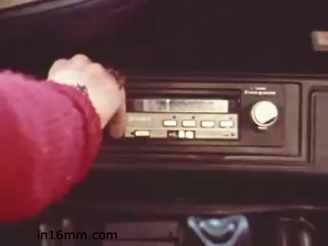 The still from the 1982 educational film 'Walk Safely' shows a close-up of a hand reaching towards a car's stereo system. The interior of the vehicle has a vintage aesthetic consistent with the early 1980s. The focus on the stereo suggests a theme related to listening or awareness while driving. This film likely aimed to promote safe pedestrian and driving practices through everyday scenarios.