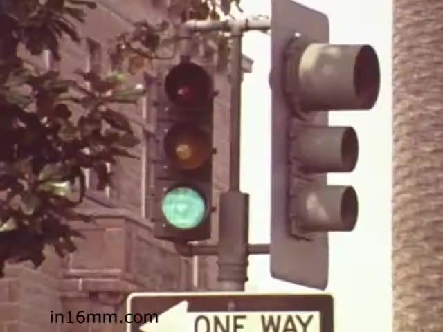 The still from 'Walk Safely - Educational Film from 1982' features a traffic light with green, yellow, and red signals visible. The green light is illuminated, indicating that vehicles are permitted to proceed. The surrounding environment includes a 'ONE WAY' sign, suggesting a focus on traffic rules and safe pedestrian practices. The context implies a teaching moment about road safety, typical of educational films from that era, aimed at promoting responsible behavior in traffic settings.
