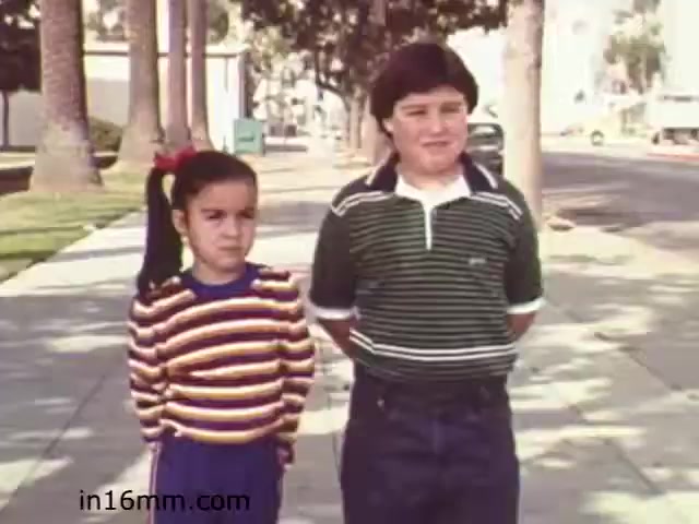 The still from 'Walk Safely,' an educational film from 1982 by Fiesta Films, features two children standing on a sidewalk. The girl on the left wears a colorful striped sweater and has a ponytail, while the boy on the right is dressed in a green and white striped shirt with navy blue pants. Palm trees and a street are visible in the background, suggesting a suburban setting. The image likely emphasizes themes of safety and awareness for children, reflective of educational content from that era.