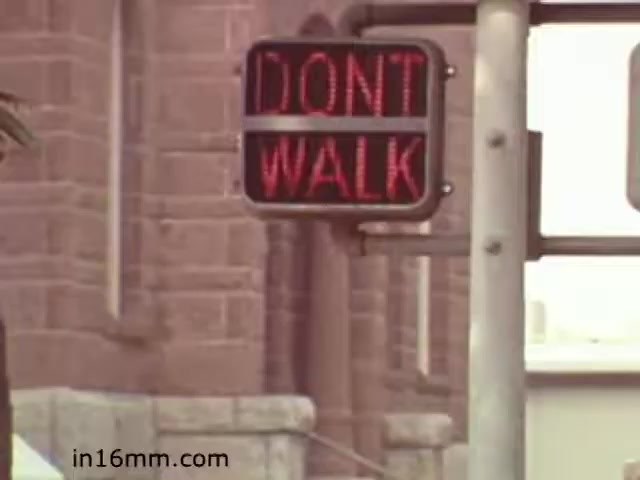 The still from 'Walk Safely,' an educational film from 1982 by Fiesta Films, features a prominent traffic signal displaying the message 'DON'T WALK.' The context suggests a focus on pedestrian safety, likely educating viewers about the importance of following traffic signals. The visual elements convey a clear, cautionary message intended to promote safe walking practices. The setting includes an urban environment, indicated by the nearby building architecture.