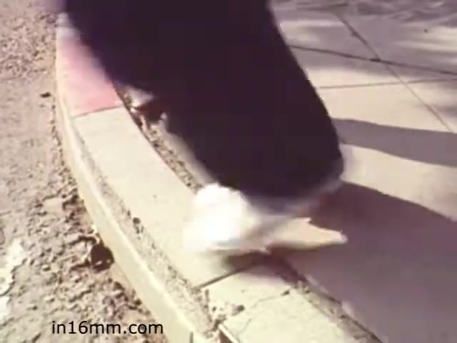 The still from 'Walk Safely' shows a close-up of feet walking on the edge of a curb. The context suggests a focus on pedestrian safety, likely illustrating the importance of walking carefully in urban environments. The film, produced by Fiesta Films in 1982, aims to educate viewers about safe walking practices. The scene emphasizes cautious movement, possibly highlighting hazards like uneven surfaces or traffic.