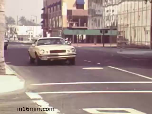 The still from 'Walk Safely,' an educational film from 1982 by Fiesta Films, shows a city street scene. A vintage yellow car is seen making a turn, indicative of the era’s automotive design. The surroundings include a mix of buildings with a somewhat dated architecture, suggesting a focus on urban environments. The film likely addresses pedestrian safety and road awareness, common themes in educational materials from that time. The overall atmosphere conveys a sense of mid-20th century urban life.