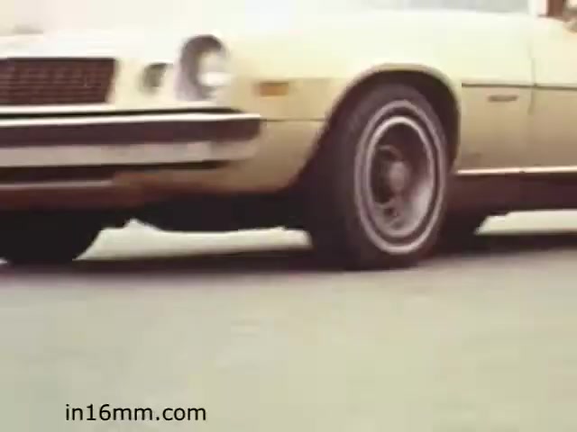 The still from 'Walk Safely,' an educational film from 1982 by Fiesta Films, features a yellow car, likely from the late 1970s or early 1980s, in motion. The focus is on the vehicle's wheel and lower body, suggesting a perspective that emphasizes driving safely. The film likely addresses themes of pedestrian safety and roadway awareness, common in educational films of that era.