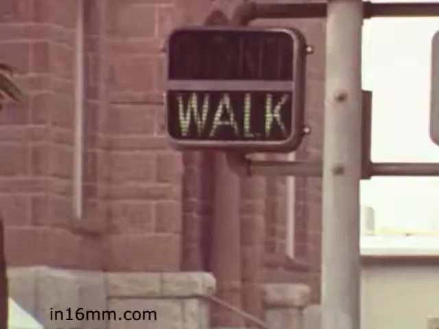 The still from 'Walk Safely - Educational Film from 1982 - Fiesta Films' features a pedestrian traffic signal displaying the word 'WALK.' The image captures a moment intended to emphasize safe walking practices, likely aimed at educating viewers about pedestrian safety. The background hints at an urban environment, with a building partially visible. This film is part of a collection of educational materials, possibly targeting both children and adults to promote awareness around walking in traffic.