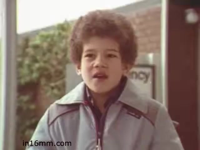 The still from 'Walk Safely,' an educational film from 1982, features a young boy with an expressive face and curly hair, dressed in a light blue jacket. The setting appears to be outdoors, possibly near a building or a street, with some greenery in the background. The film likely focuses on teaching children about safety while walking, reflecting the educational tone of that era. The specific emotional expression of the boy suggests he may be engaged in a discussion or learning moment.