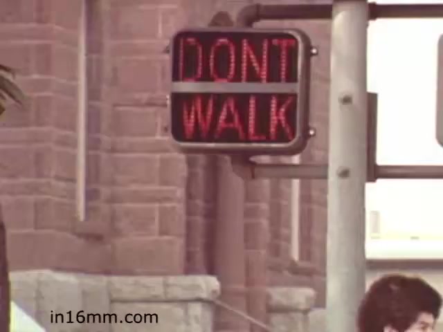The still from 'Walk Safely,' an educational film from 1982 by Fiesta Films, features a traffic signal displaying the message 'DON'T WALK.' This sign is intended to instruct pedestrians to wait before crossing the street. The setting suggests an urban environment, emphasizing road safety themes typical of educational films aimed at teaching safe walking practices.