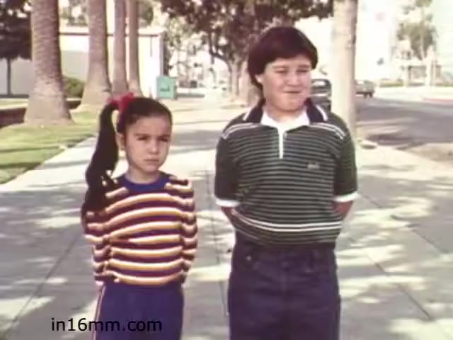 The still from the 1982 educational film 'Walk Safely' features two children standing together on a sidewalk lined with palm trees. The girl on the left wears a brightly colored striped sweater and has her hair tied in a ponytail. She looks serious, while the boy on the right wears a striped polo shirt and appears more relaxed. The background includes a city street and trees, giving a sense of an urban environment. The scene likely emphasizes pedestrian safety themes, typical for educational films of that era.