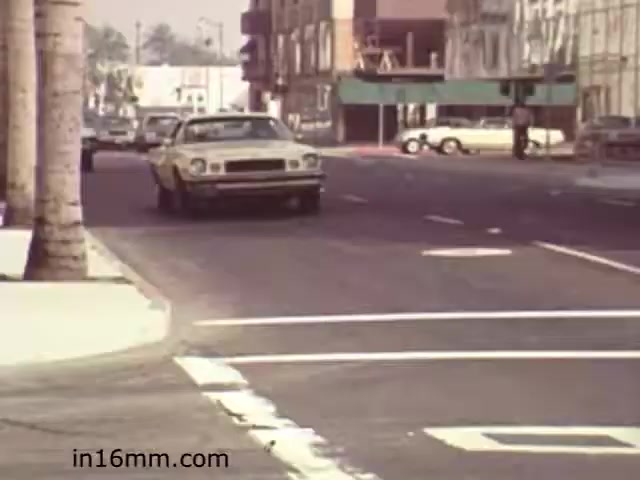 The still from 'Walk Safely,' an educational film from 1982 by Fiesta Films, depicts a street scene featuring a yellow car driving down an urban road. The setting includes palm trees, nearby buildings, and other vehicles parked along the side of the road. The image conveys a typical early '80s suburban environment, highlighting road safety themes likely explored in the film. The composition suggests a focus on pedestrian awareness and safe navigation in traffic.