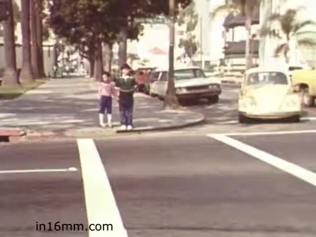 The still from 'Walk Safely,' an educational film from 1982, depicts two children standing at the curb, preparing to cross a street. They are holding hands, suggesting they are about to cross together. The scene includes a well-maintained urban environment with palm trees lining the sidewalk and parked cars along the street, indicative of the era. The use of vibrant colors reflects the style of the time, and the crossing lines are clearly marked on the road, emphasizing the film's focus on pedestrian safety.