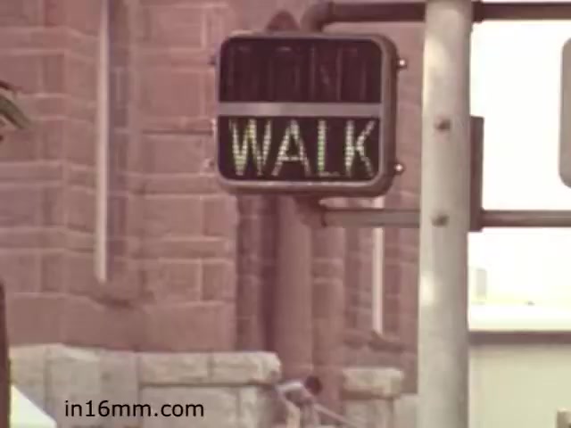 The still from 'Walk Safely,' an educational film from 1982 by Fiesta Films, features a pedestrian traffic signal displaying the word 'WALK.' This image likely emphasizes safe walking practices, targeting pedestrian awareness in urban settings. The backdrop suggests a city environment, possibly aiming to educate viewers about road safety and traffic signals. The aesthetic is characteristic of early '80s educational films, often utilizing straightforward visuals to convey important messages.