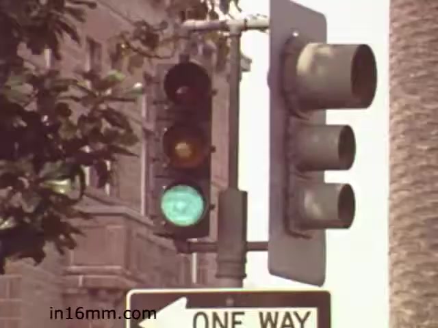 The still shows a traffic light with a green light illuminated, signaling that it is safe to proceed. Next to the traffic light is a 'ONE WAY' street sign, indicating the direction vehicles should travel. The background features a blurred cityscape, suggestive of an urban environment, along with foliage from nearby trees. The overall aesthetic is typical of educational films from the early 1980s, capturing a moment focused on pedestrian safety and traffic regulations.