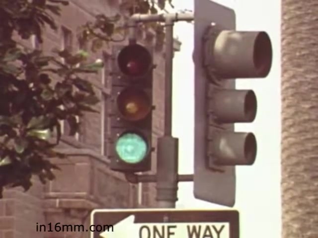 The still from 'Walk Safely - Educational Film from 1982' features a traffic light displaying a green signal, indicating that it is safe for pedestrians to cross. The setting includes a 'One Way' street sign, suggesting a focus on traffic rules and pedestrian safety. The image likely serves as a visual aid to instruct viewers on safe walking practices around traffic intersections. The style and quality reflect the educational film era of the early 1980s.