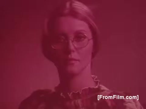 The still from 'Life Savers Gum - 1970's' features a woman with a serious expression, wearing round glasses and a patterned outfit. The image has a reddish tint, enhancing the retro aesthetic typical of that era. The background is simple and complements her look, focusing attention on her face. This captures the nostalgic and playful spirit of 1970s advertising.