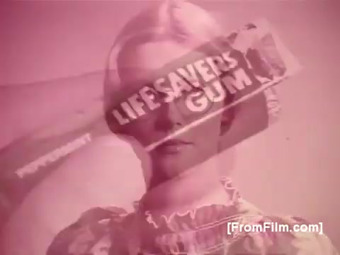 The still from the 1970s Life Savers Gum advertisement features a vintage aesthetic, with a soft pink hue dominating the image. A woman with a classic hairstyle is partially obscured by a prominent display of Life Savers Gum packaging. The design showcases the gum's branding and evokes nostalgia for the era, emphasizing the product's appeal with a playful and lighthearted tone.