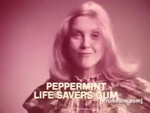 The still features a woman with long, wavy hair, smiling at the camera. She is dressed in a patterned outfit, exuding a retro 1970s vibe. Text at the bottom highlights 'PEPPERMINT LIFE SAVERS GUM' in bold, white letters against a soft, pink background, emphasizing the product. The overall aesthetic reflects the playful and vibrant advertising style of that era.