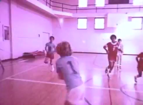 The still from 'Do it for them - take your blood pressure medication 5' features a gymnasium scene where children are engaged in a basketball game. Some wear blue uniforms while others are in red, capturing a lively and active atmosphere. The gym is well-lit, emphasizing the importance of physical activity and teamwork, reinforcing the message about health and taking medication. The color tone appears warm and slightly faded, typical of 16mm film.