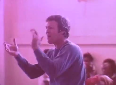 The still depicts a man in a blue sweater, animatedly gesturing as he speaks, possibly conveying an important message. The background features blurred figures, suggesting a group setting, likely indicating engagement or participation. The overall scene has a pinkish hue, contributing to a vibrant yet serious atmosphere related to health awareness, specifically about taking blood pressure medication.