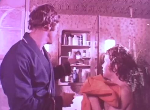 The still features two individuals in a bathroom setting. One person, dressed in a robe, is turning towards a child or young person, who is wrapped in a towel. The adult appears to be reaching for something from a cabinet filled with various medication bottles. The overall atmosphere conveys a moment of care and attention, likely emphasizing the importance of taking blood pressure medication. The color palette has a soft, vintage tone characteristic of 16mm film.