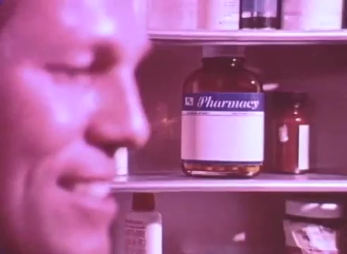 The still features a close-up of a smiling man, partially in focus, with his face illuminated against a backdrop of a medicine cabinet. Prominent in the cabinet is a large bottle labeled 'Pharmacy,' along with various smaller containers. The warm, muted colors create a reassuring atmosphere, emphasizing a theme of health and the importance of taking blood pressure medication for loved ones. The context suggests a message about personal health responsibility.