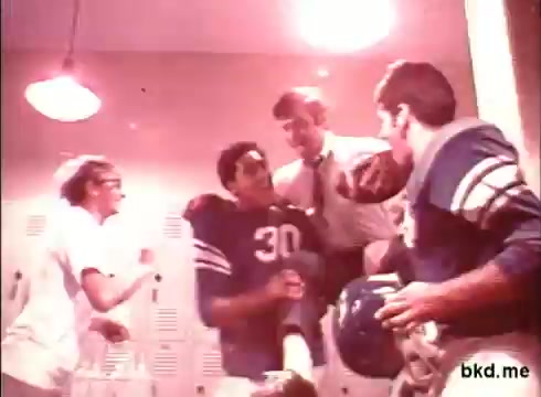 The still features a locker room scene with a mix of excitement and camaraderie. A football player in a blue jersey with the number 30 is being helped by a man in a suit, while another player in similar attire looks on, holding a helmet. A woman in a white uniform is also present, possibly a trainer. The lighting is warm and somewhat nostalgic, characteristic of 1970s commercial aesthetics.
