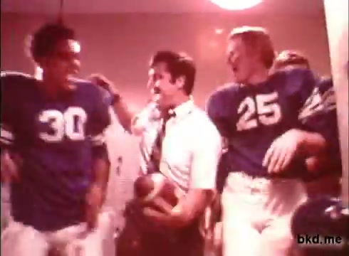 The still depicts a celebratory scene from a 1970s Crest toothpaste commercial. It features three male football players in blue jerseys, laughing and interacting with a man in a white shirt, likely a coach. The setting appears to be a locker room, illuminated with warm tones, capturing a moment of camaraderie and joy following a game. The players exhibit a spirited and energetic atmosphere, emphasizing a youthful and fun portrayal of dental care.