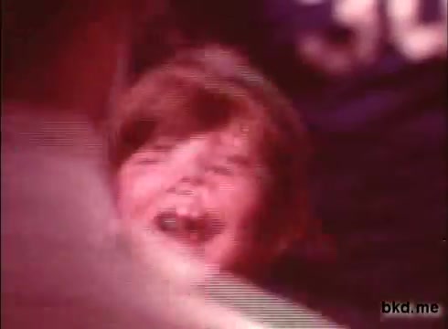 The still features a young child joyfully laughing, likely in a playful or celebratory context. The image has a vintage look characteristic of 16mm film, with warm tones and some graininess. There are indistinct figures in the background, enhancing the lively atmosphere typical of 1970s commercials.