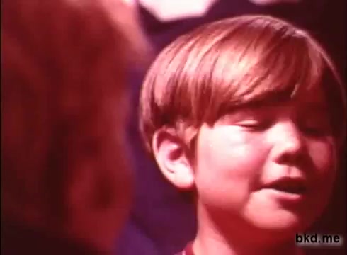 The still from the 1970s Crest toothpaste commercial shows a young boy with short, light brown hair, appearing engaged and animated as he talks or sings. The background features soft, blurred colors that suggest a playful atmosphere. The image has a warm, nostalgic tone typical of vintage advertisements from that era.