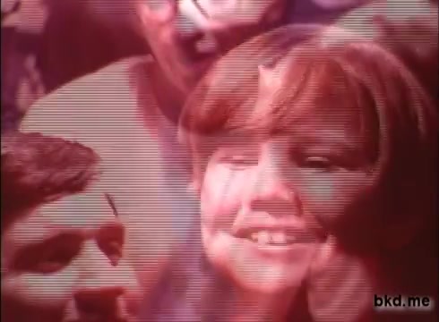 The still from the 1970s Crest toothpaste commercial features a vibrant, colorful composition with a close-up of a smiling girl in the foreground. Her joyful expression contrasts with blurred faces of adults in the background, creating a nostalgic and lively atmosphere. The overall tone is cheerful, emphasizing the positive effects of the product, reflective of the era's advertising style.